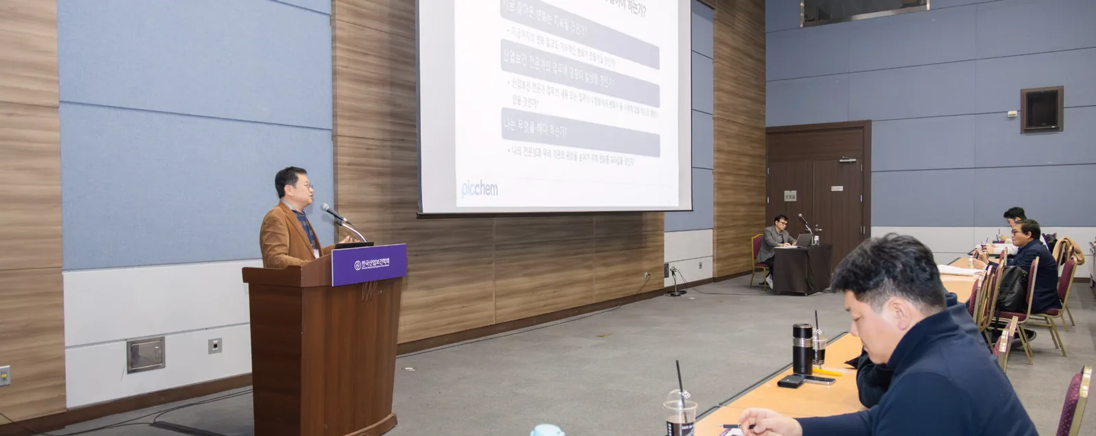 PDC2 두 번째 강의 현장 — 한국산업보건학회 연단에서 강의하는 픽켐 김신범 대표와 스크린에 띄운 픽켐 발표 자료(‘지금 찾아온 변화는 지속될 것인가?’, ‘나는 무엇을 해야 하는가?’ 등 세 가지 질문), 책상에 앉아 듣는 수강생들