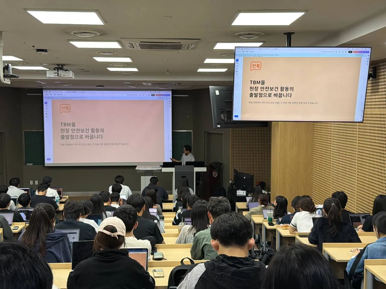고려대학교 특강 현장 — 강단의 정태진 대표와 스크린·모니터에 띄운 안톡 슬라이드 ‘TBM을 현장 안전보건 활동의 출발점으로 바꿉니다’, 뒷모습의 수강생들